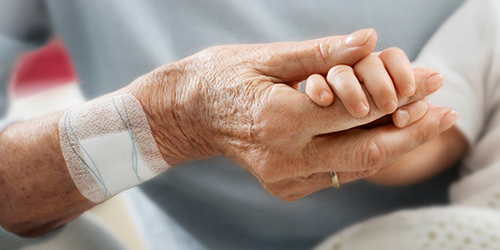 En ældre kvinde med et sensitive plaster på højre arm holder et spædbarns hånd.  En ældre kvinde med et sensitive plaster på højre arm holder et spædbarns hånd.